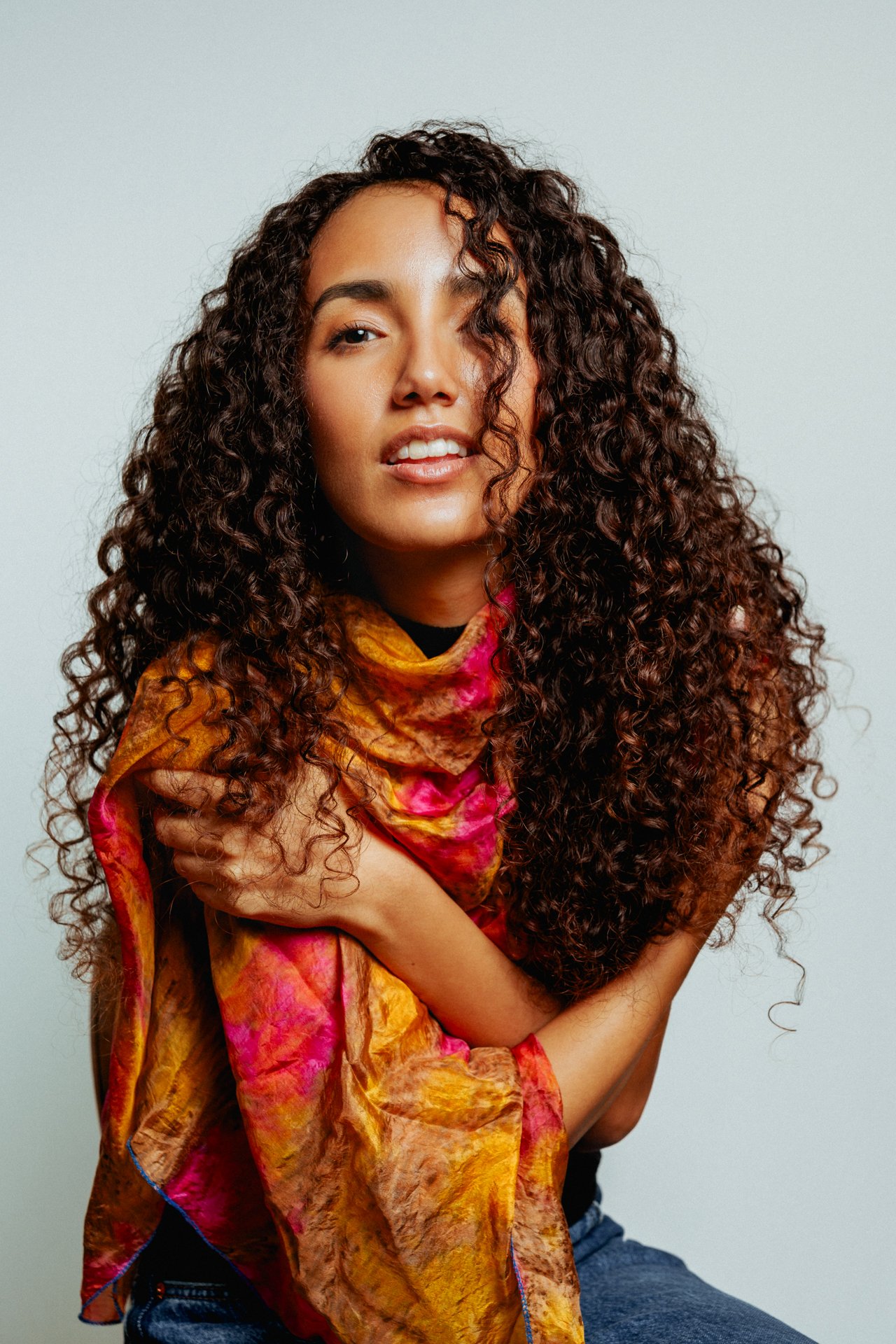 Donna con grandi capelli ricci e sciarpa colorata seduta contro sfondo bianco.