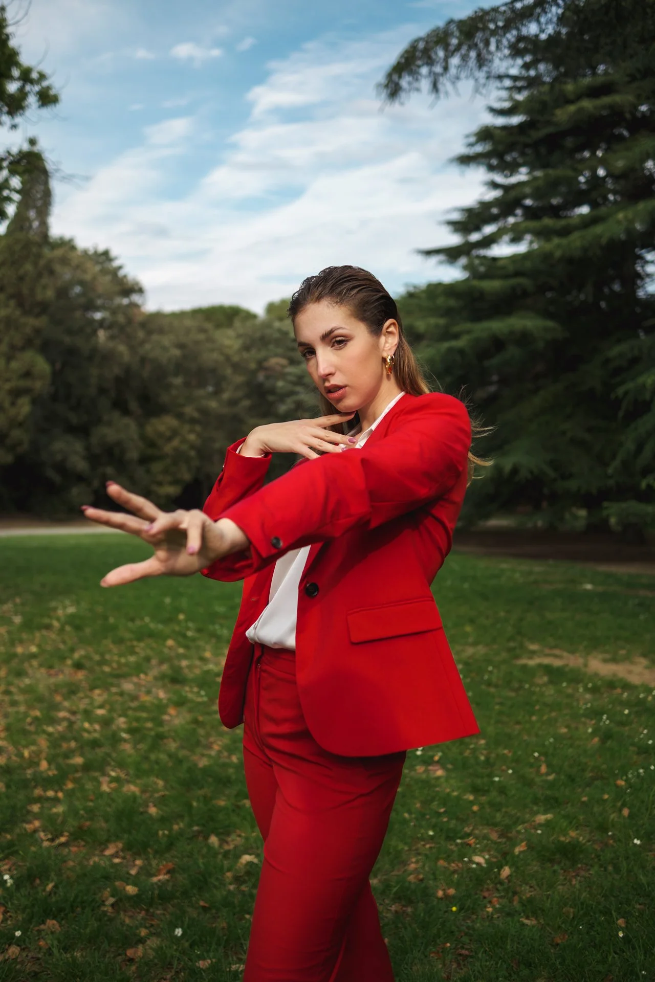 Una donna in giacca e pantaloni rossi posa in un parco, con uno sfondo di alberi e cielo.