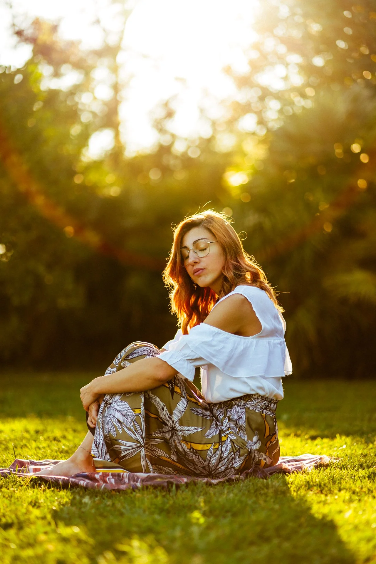 Ragazza con capelli rossi, vestita con pantaloni leggeri con fantasia, seduta sull'erba in un giardino durante il tramonto.