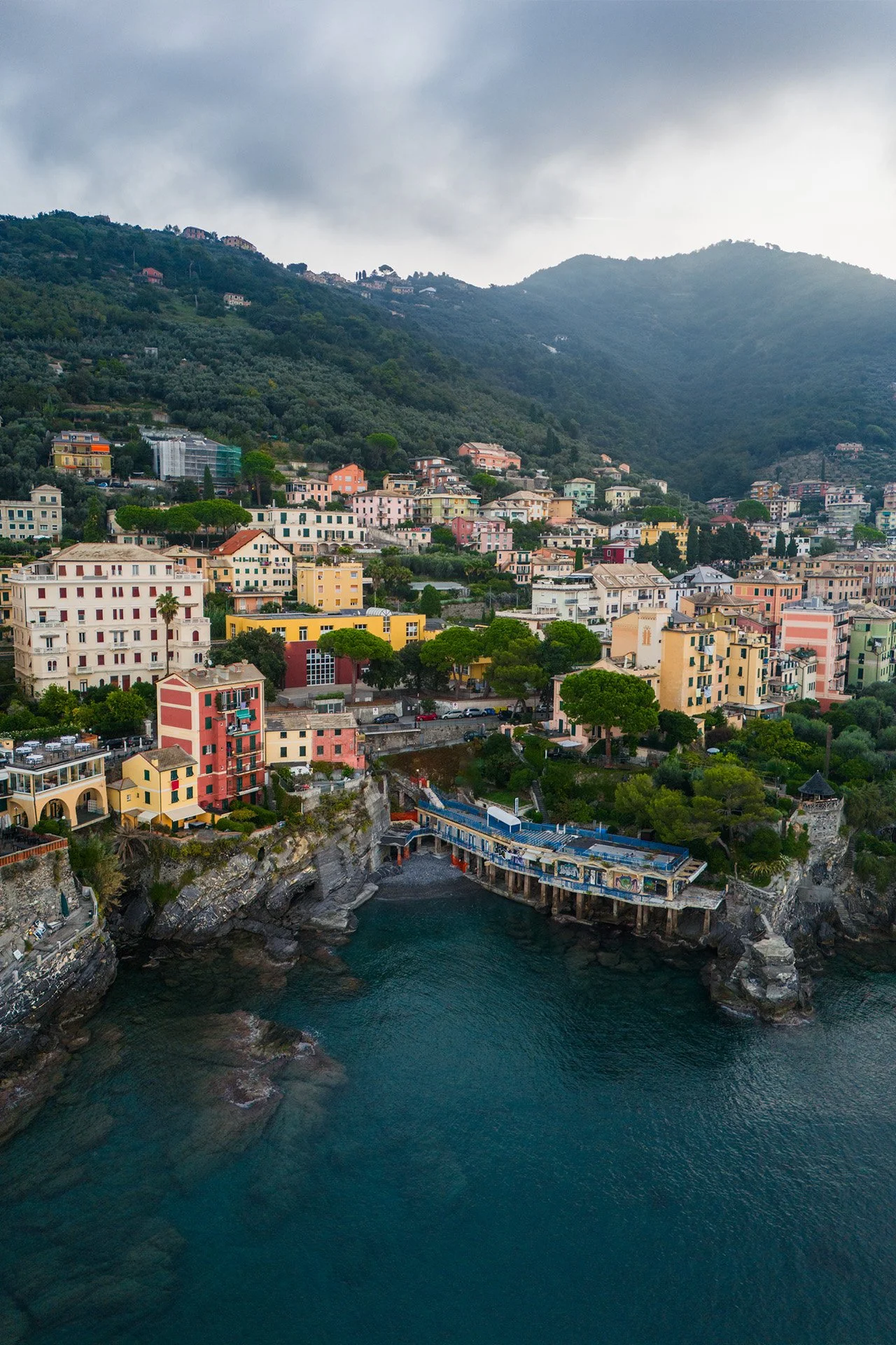 Paesaggio costiero con case colorate su una scogliera, mare blu, e colline verdi con nuvole grigie.