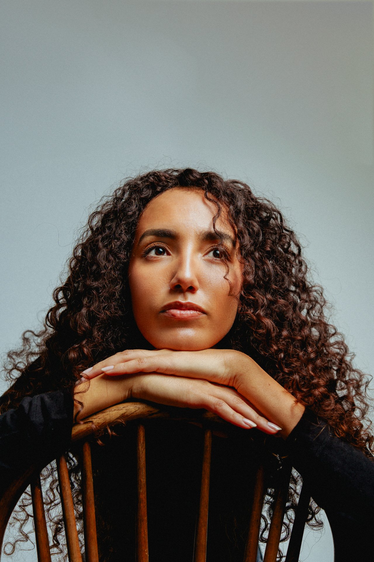 Ragazza dai capelli riccioli appoggiata con il mento sulle mani, seduta su una sedia in legno, sfondo neutro