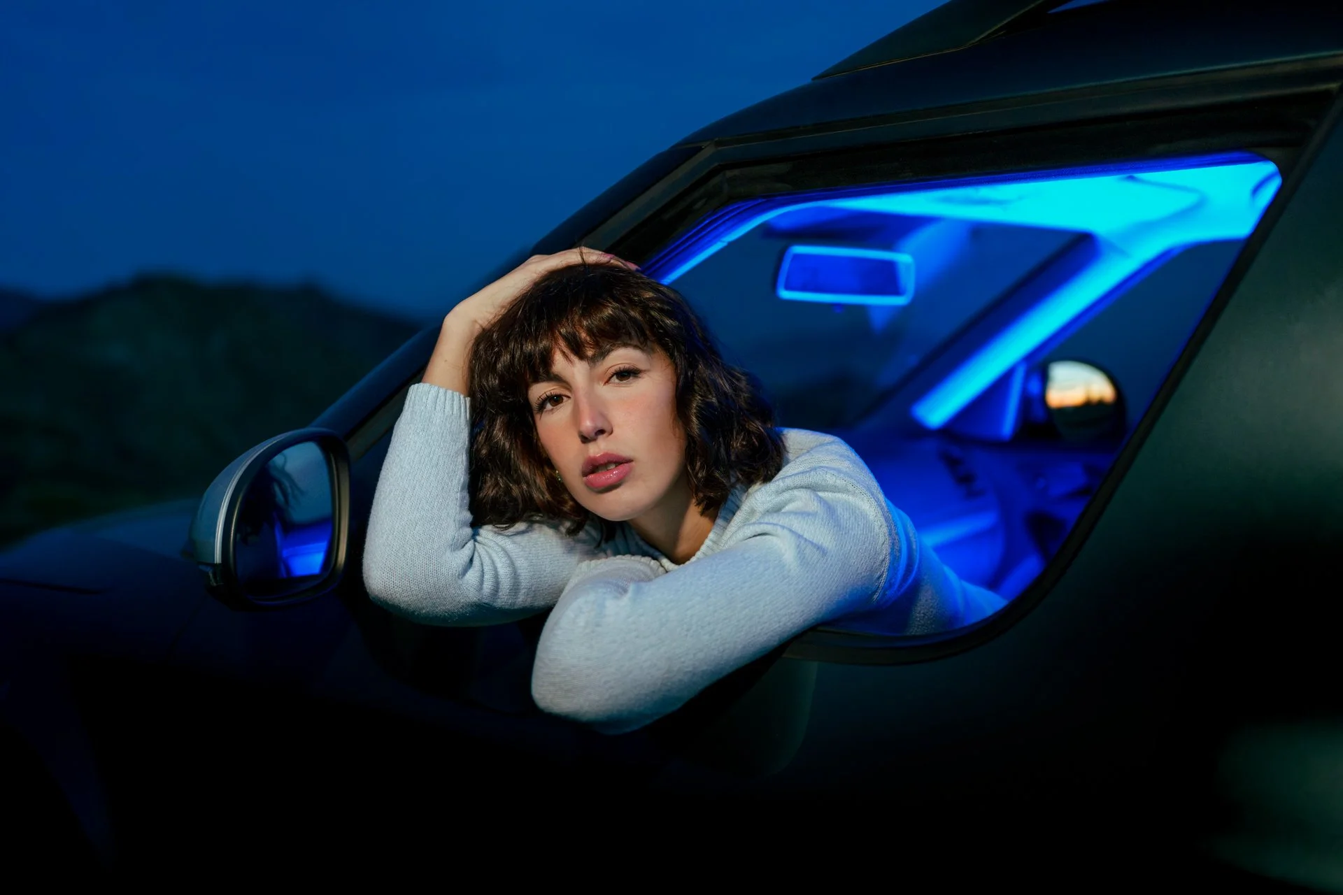 Donna che posa con la testa appoggiata sul braccio nel finestrino di un'auto con luci blu al interno, di sera o notte.