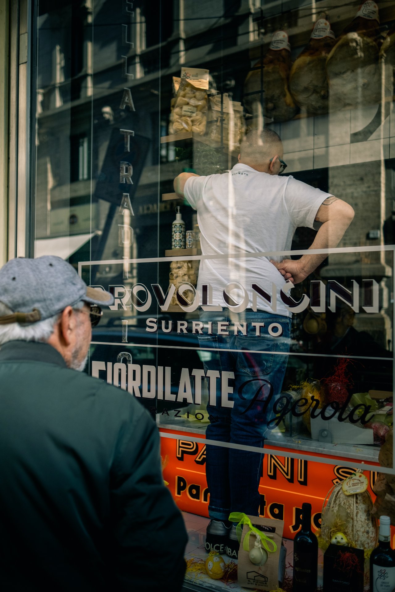 Due uomini davanti a una vetrina di una gastronomia, uno di loro con cappello e occhiali, mentre l'altro, vestito di bianco, sta preparando qualcosa dentro il negozio. La vetrina mostra vari prodotti alimentari e alcune bottiglie.