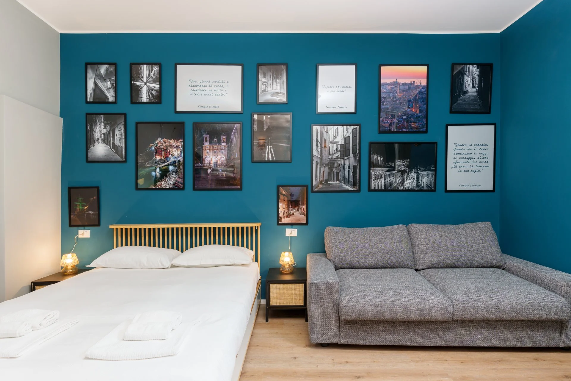 Camera da letto con parete blu decorata con una serie di quadri e foto, letto matrimoniale con lenzuola bianche e due cuscini, lampada da notte, e un divano grigio.