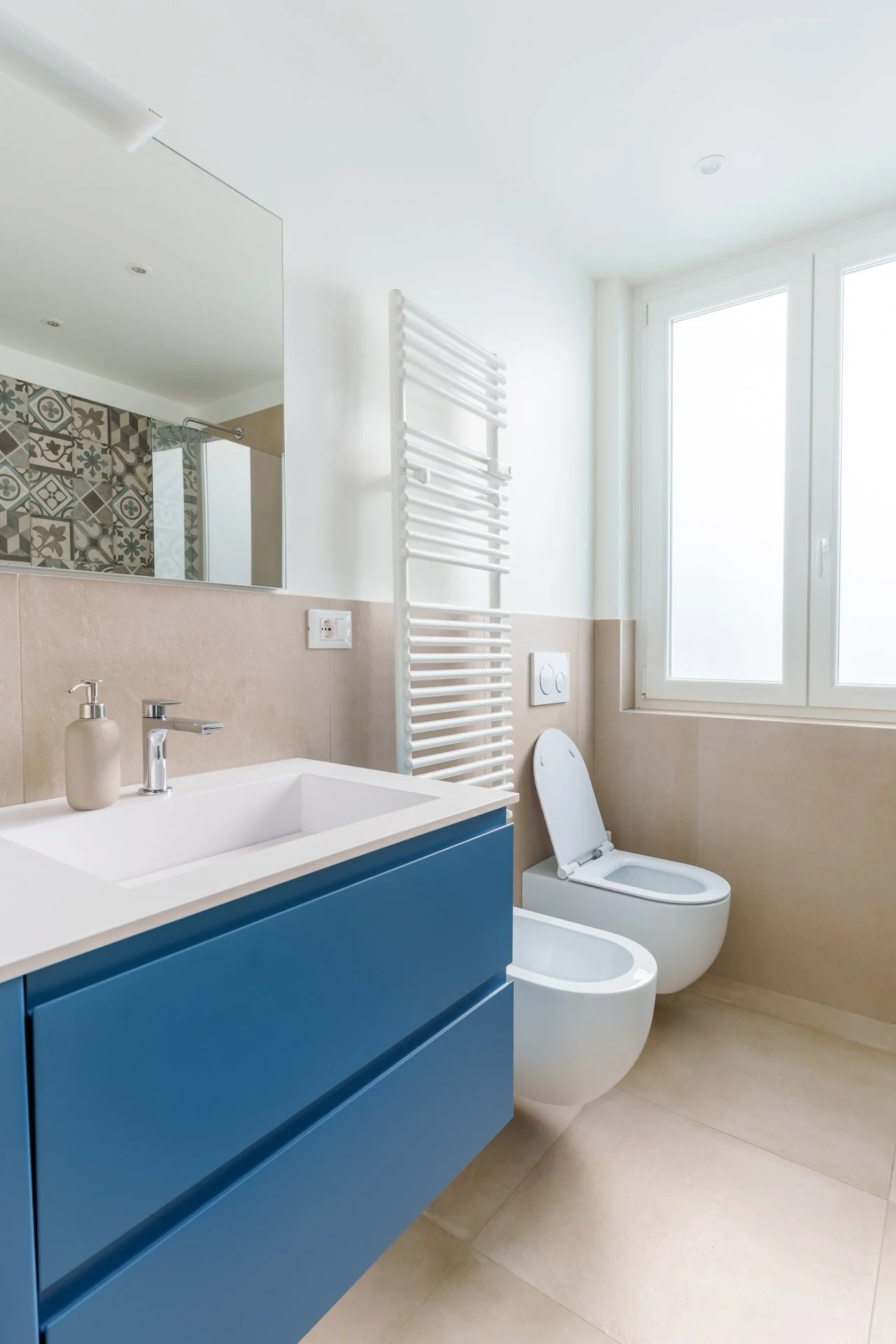 Bagno moderno con lavabo blu, specchio grande, bidet e WC bianco, finestra grande, asciugamano riscaldato bianco e pareti chiare.