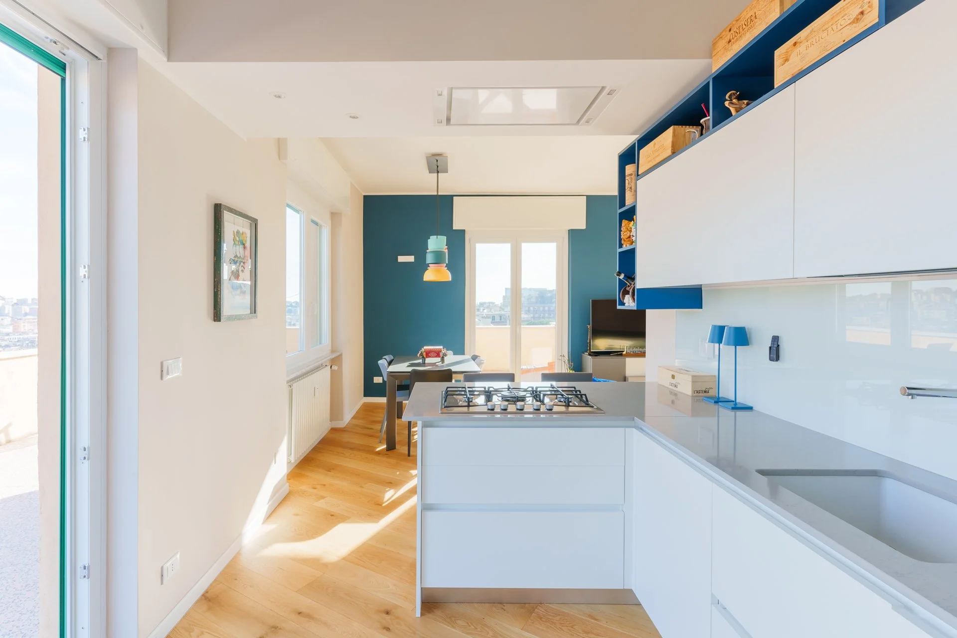 Appartamento moderno con cucina bianca, tigli in legno, pareti blu, soggiorno con TV, tavolo da pranzo con sedie, balcone con vista sulla città.