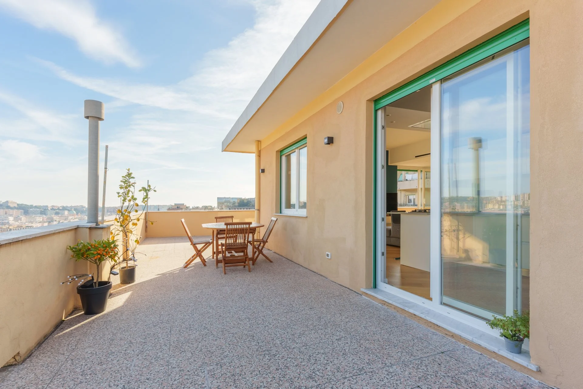 Terrazza con tavolo e sedie in legno, piante in vasi, apertura di porta scorrevole e vista sulla città.