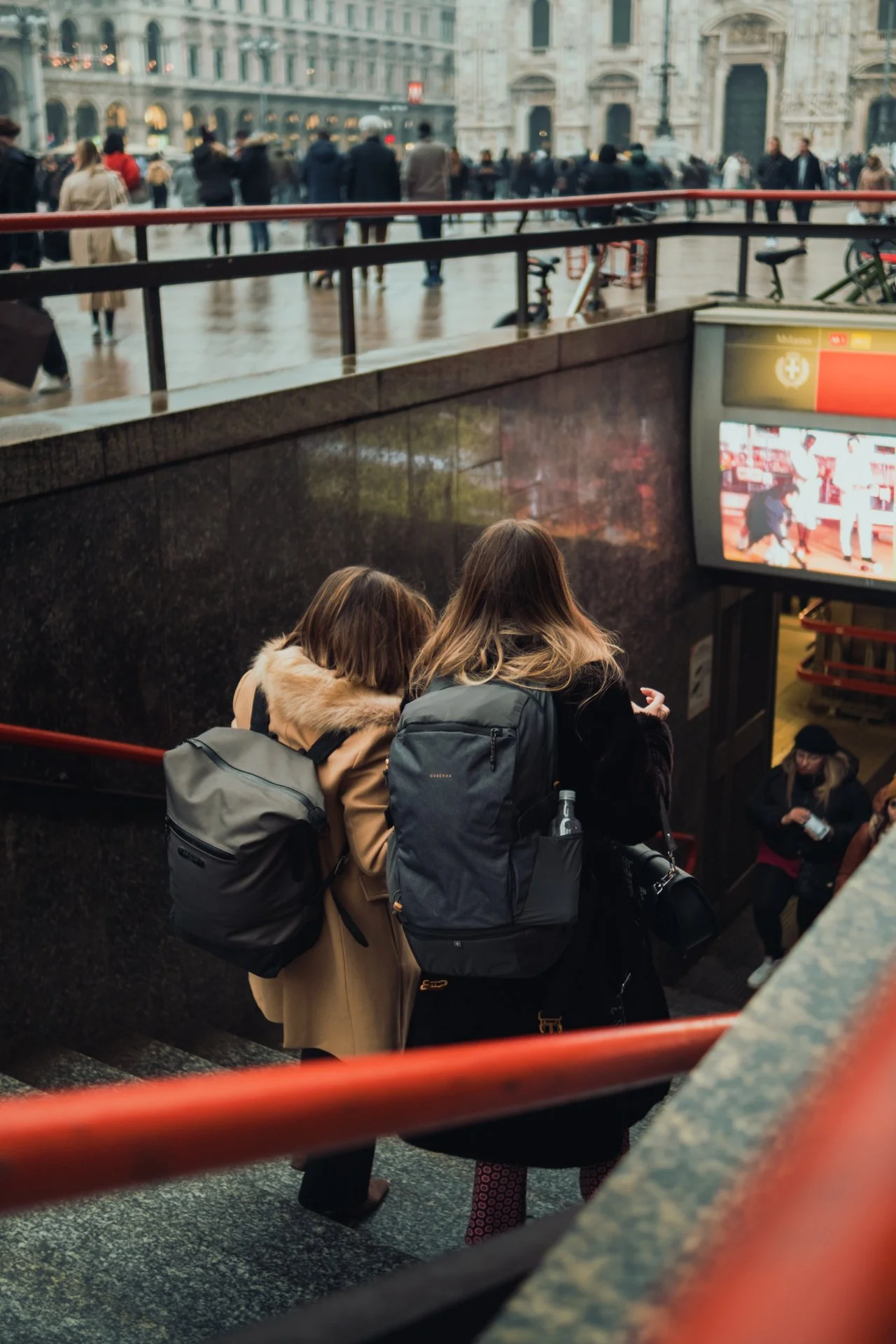 Due donne con zaini in piedi sulla salita di una metropolitana, con una scena urbana affollata sullo sfondo.