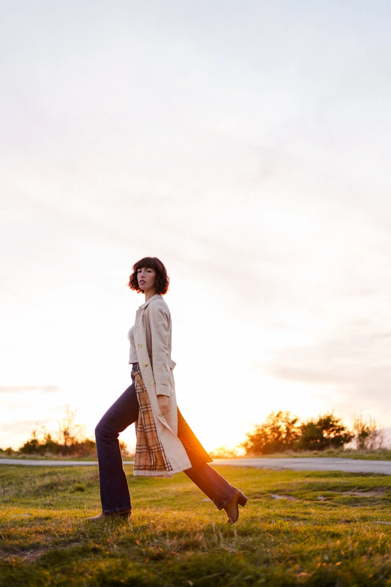 Una donna che cammina all'aperto durante il tramonto con un trench beige, pantaloni larghi a quadri e scarpe con tacco, sullo sfondo un cielo nuvoloso e alberi lontani.