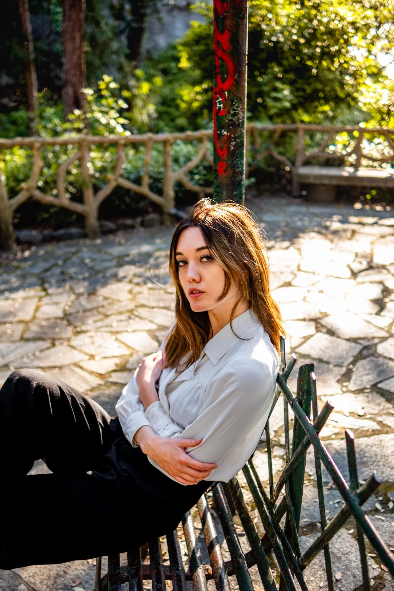 Ragazza seduta su una panchina in un parco, con abbigliamento bianco e pantaloni neri, con sfondo di alberi e una recinzione in metallo