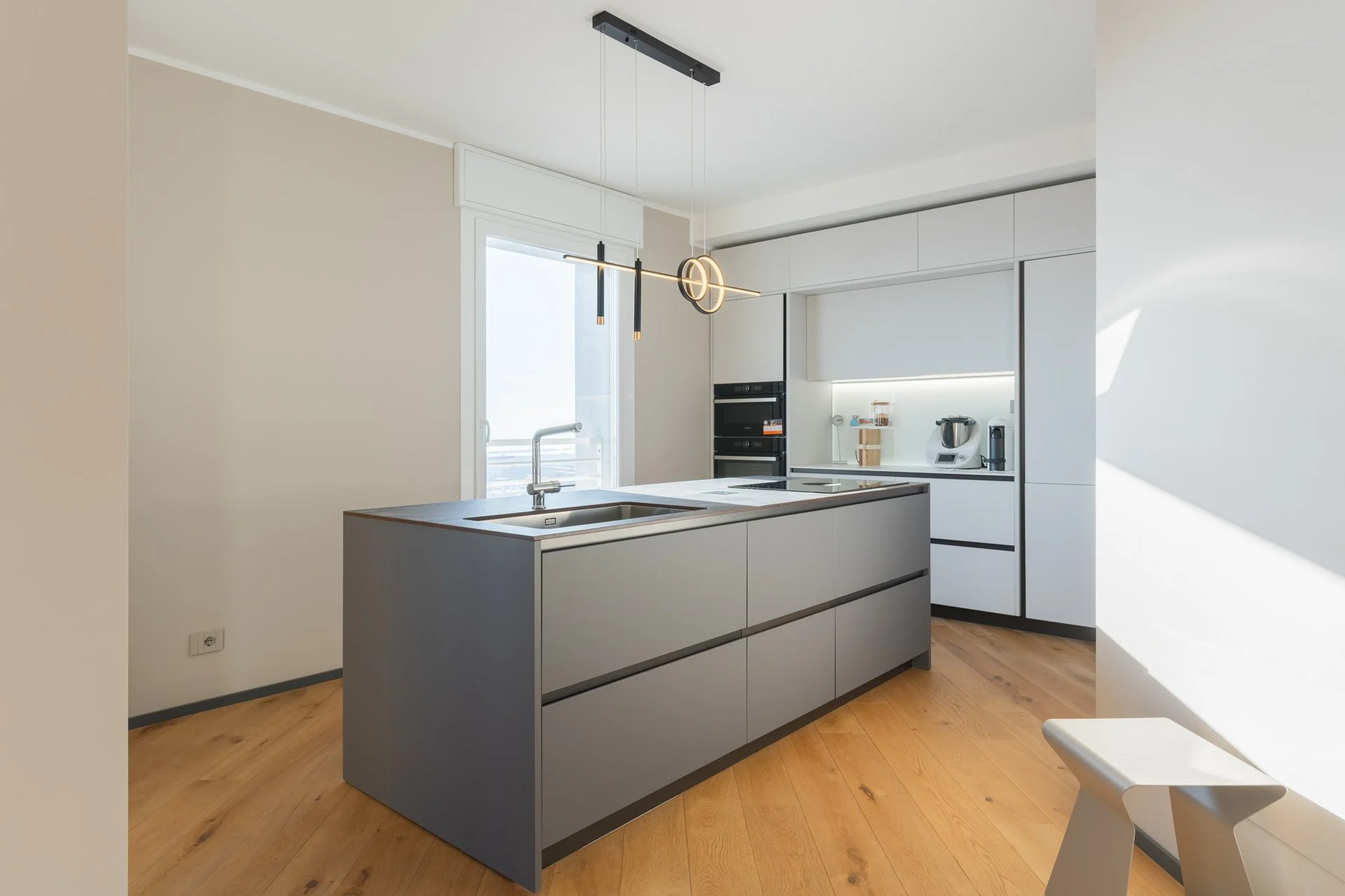 Cucina moderna bianca con isola grigia, luci pendenti e armadietti in bianco, pavimento in legno e porta finestra con balcone.