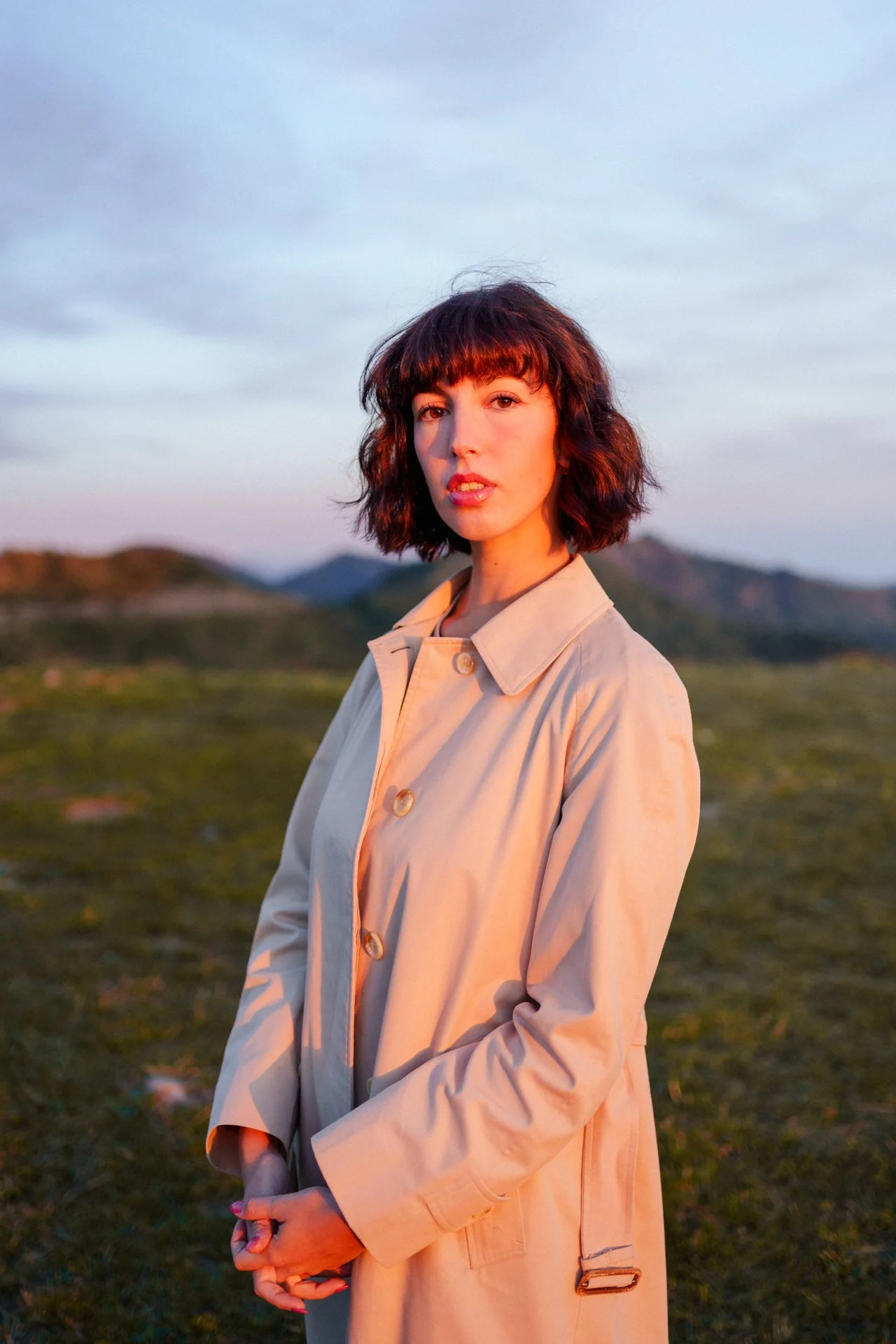 Una donna con capelli ricci castani chiari indossa un trench beige in un paesaggio naturale al tramonto, con montagne e cielo nuvoloso sullo sfondo.