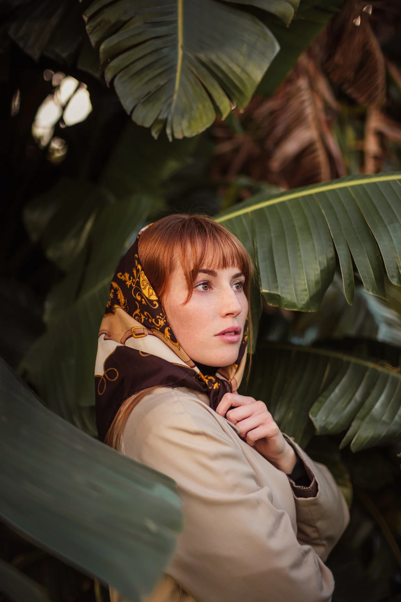 Femme aux cheveux roux portant un foulard coloré, entourée de grandes feuilles vertes tropicales, regarde pensivement à droite.