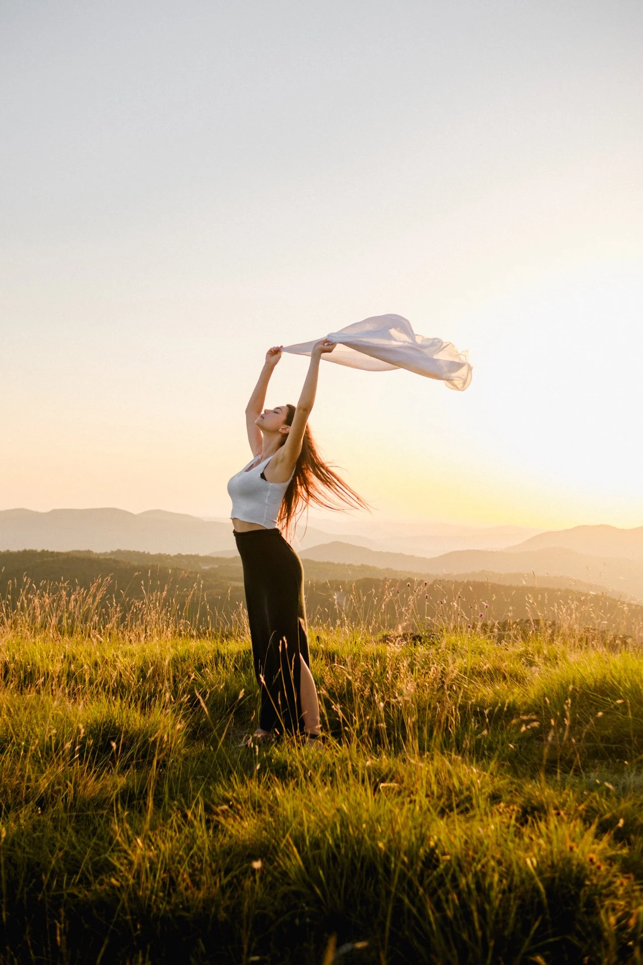 Una donna si trova in un campo erboso durante il tramonto, tiene un tessuto bianco in aria con entrambe le mani, indossa un top bianco e una gonna nera con spacco.