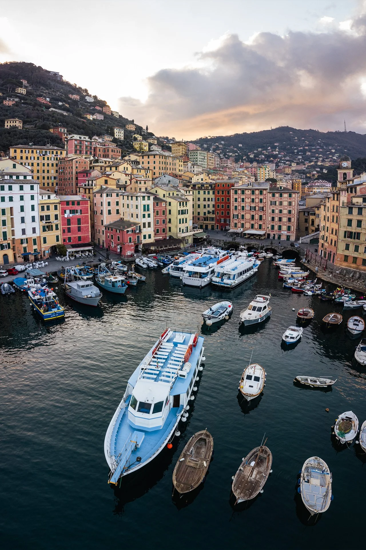 Un porto con barche e yacht ormeggiati, con edifici colorati e case che salgono lungo la collina, sotto un cielo nuvoloso al tramonto.