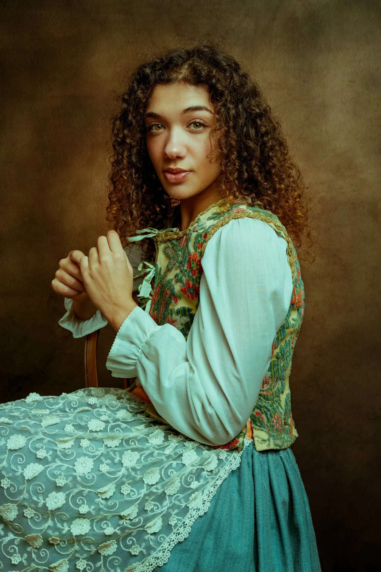 Una donna con capelli ricci, vestita con abiti tradizionali, seduta contro uno sfondo marrone, che sorride leggermente.