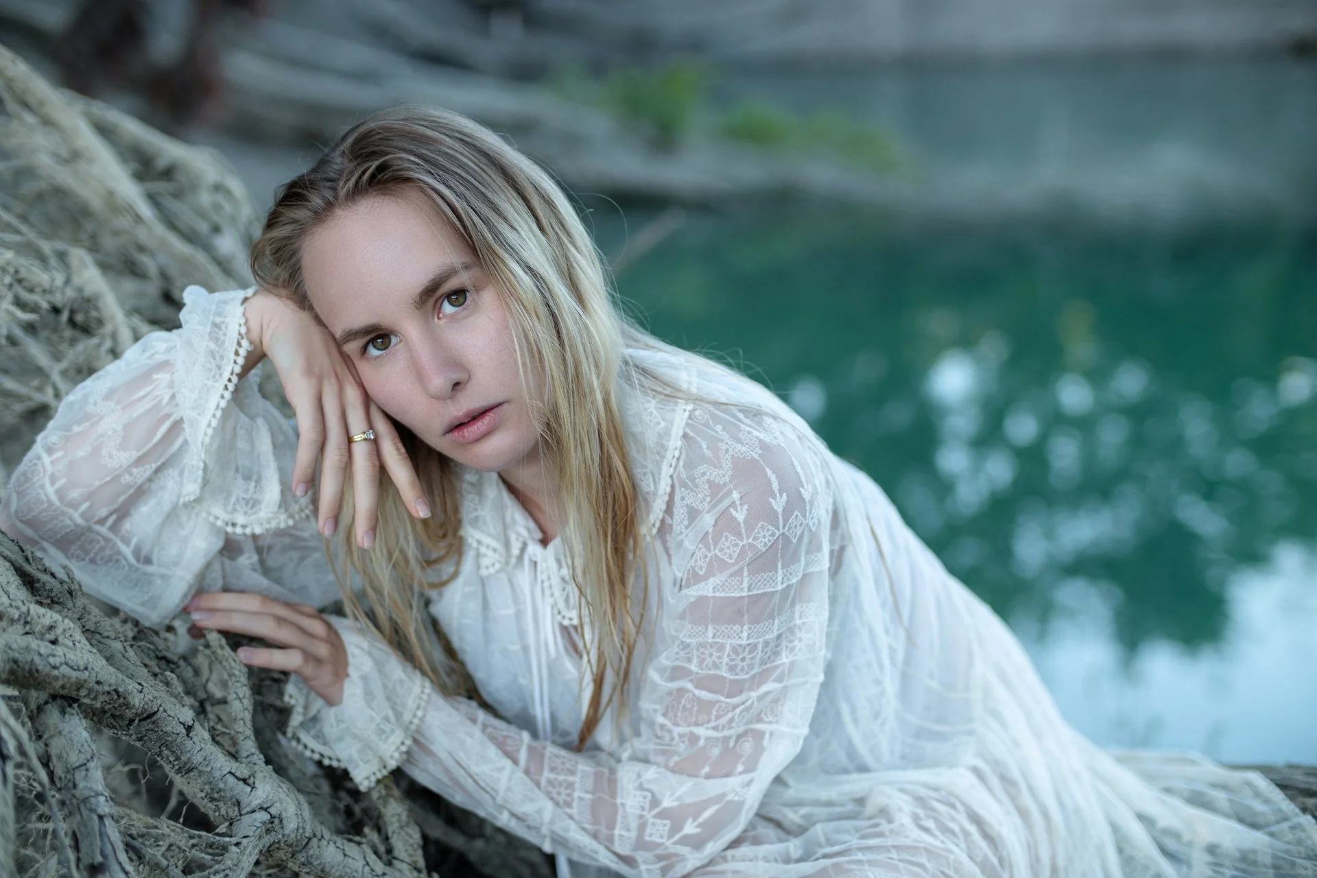 Una donna con capelli biondi chiari e occhi azzurri giace su un tronco vicino all'acqua, indossando un vestito bianco in pizzo con dettagli ricamati.