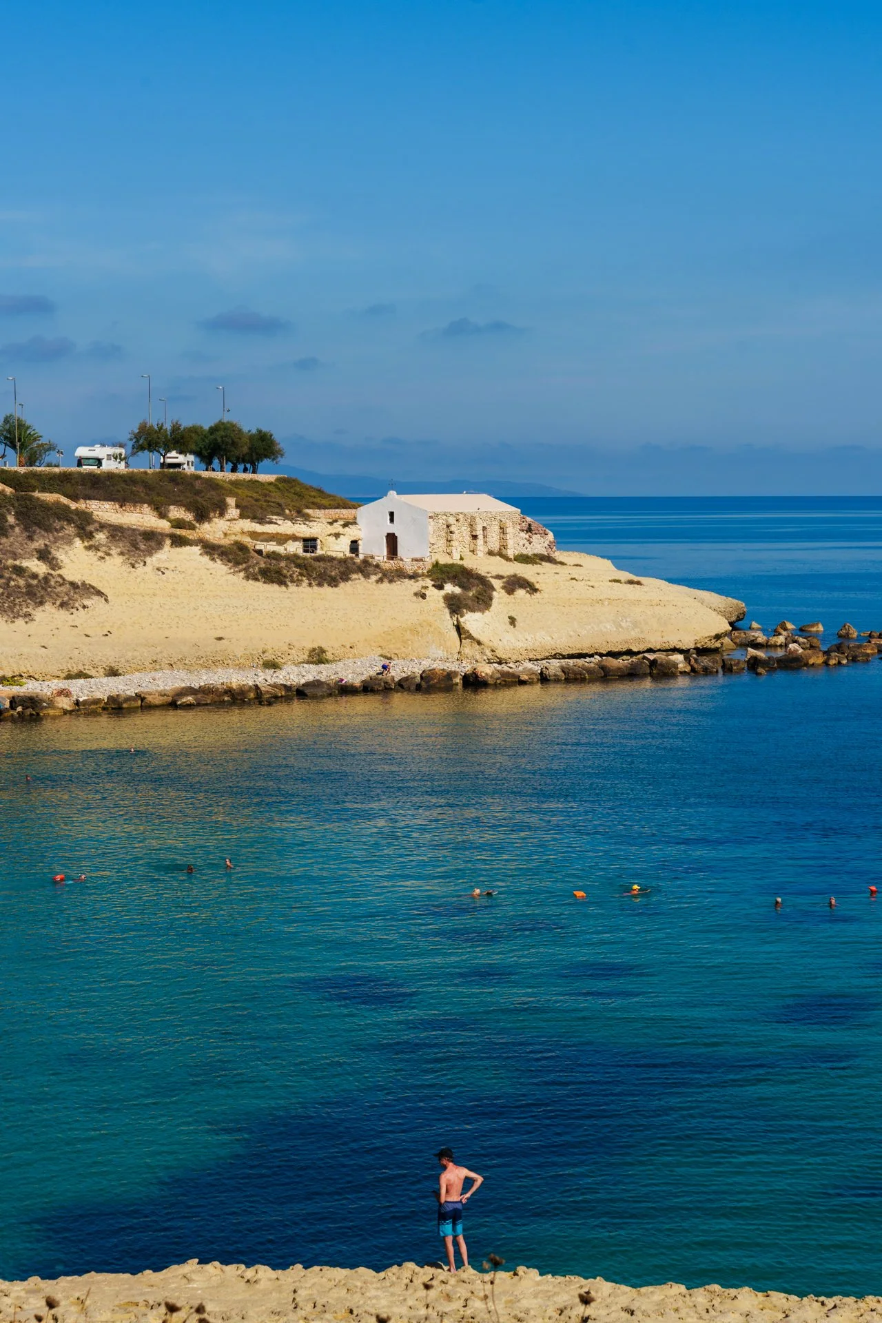Spiaggia con un uomo in costume da bagno che guarda il mare, acque tranquille, scogliera con piccola chiesa bianca, e alcune persone che nuotano o si rilassano in acqua. Sul protagonista, scena di un ambiente di mare durante giornate soleggiate.