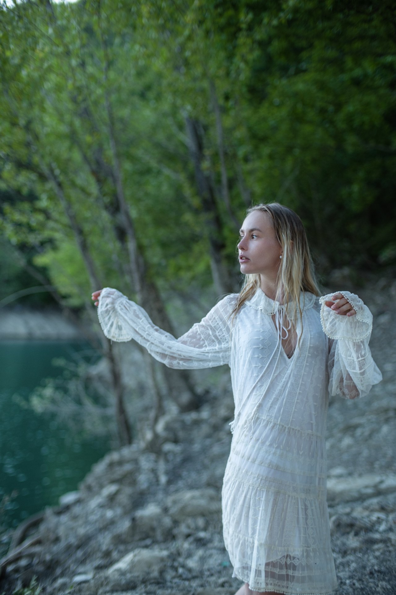 Donna con abito bianco lungo, capelli biondi, in un ambiente naturale con alberi e acqua, sembra in movimento.