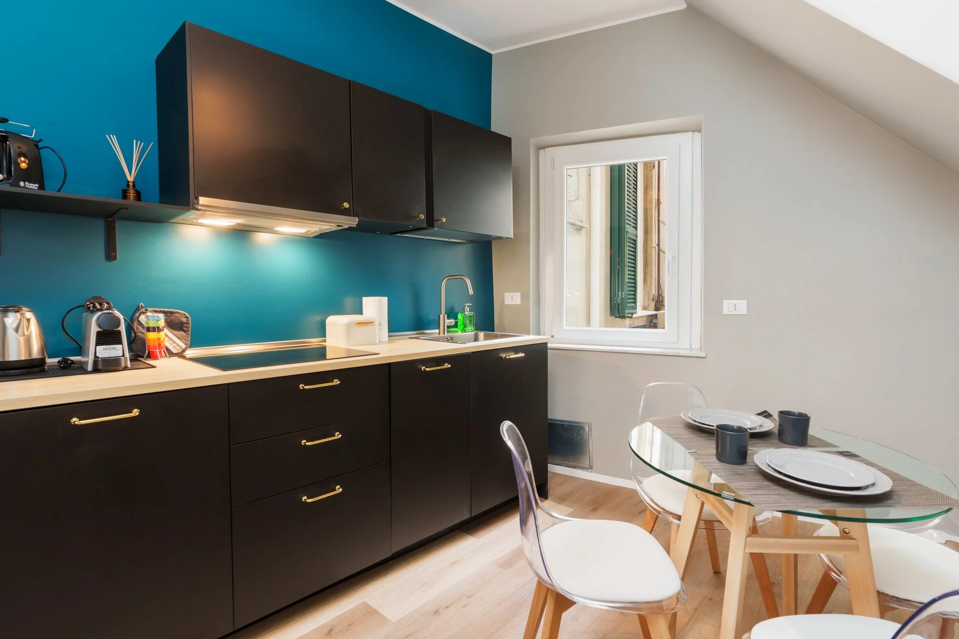 Cucina moderna con credenze nere, pareti blu e beige, tavolo rotondo con piatti e bicchieri, sedie trasparenti, finestra con serrature verdi.