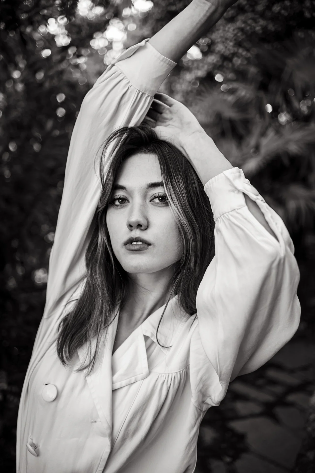 Ragazza con capelli lunghi, vestita con una camicia ampia, posa con un braccio alzato sopra la testa, sfondo di foglie sfocate.