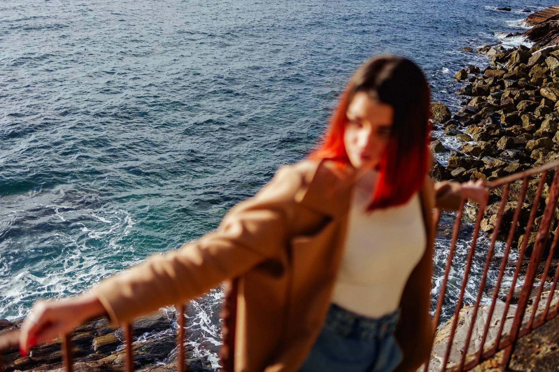 Una donna con i capelli rossicci e con una gonna bianca, che si appoggia a una ringhiera vicino al mare, con onde che si infrangono contro le rocce.