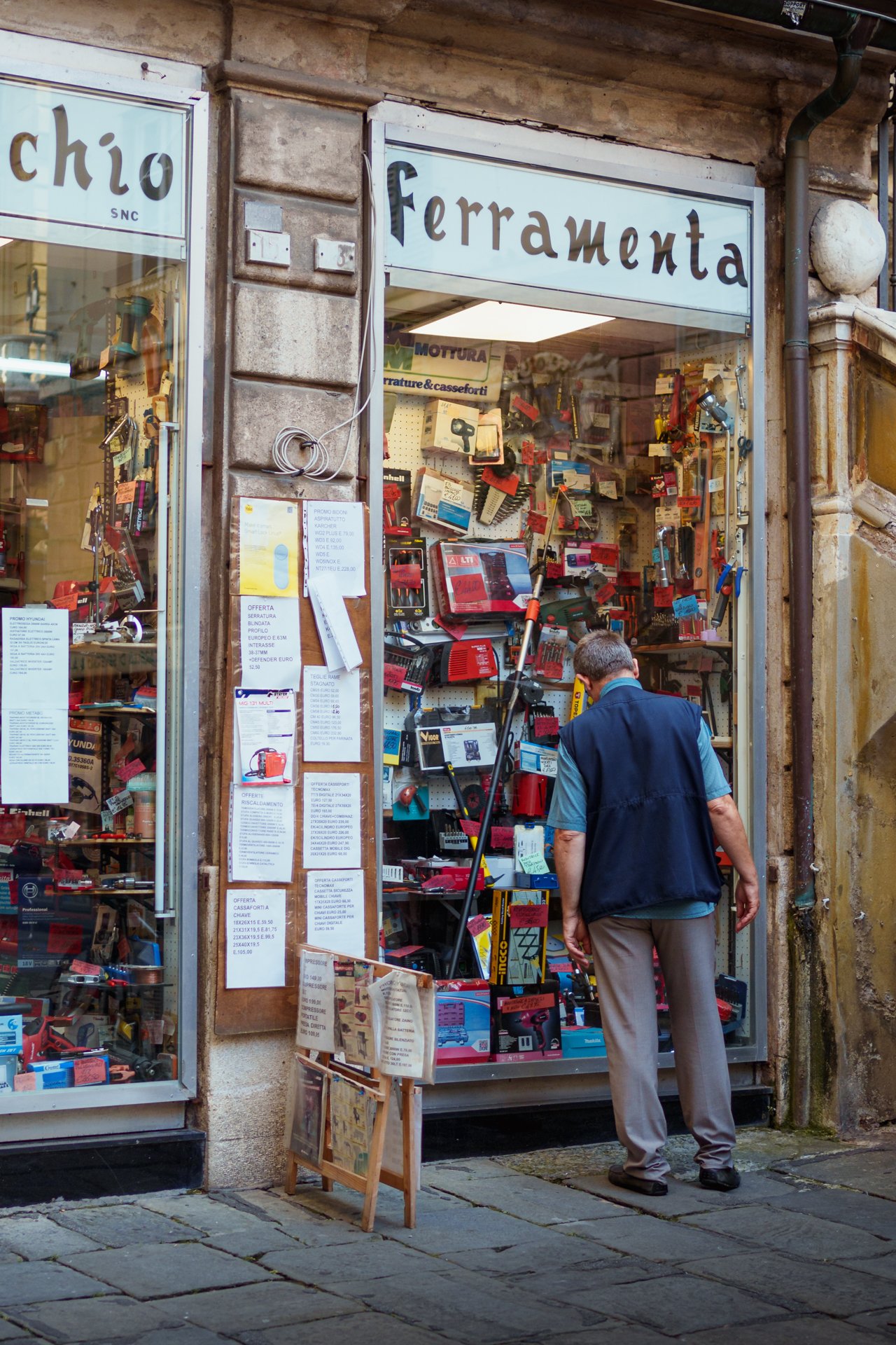 Un uomo che osserva strumenti da ferramenta in un negozio sui ciottoli di una strada di pietra.