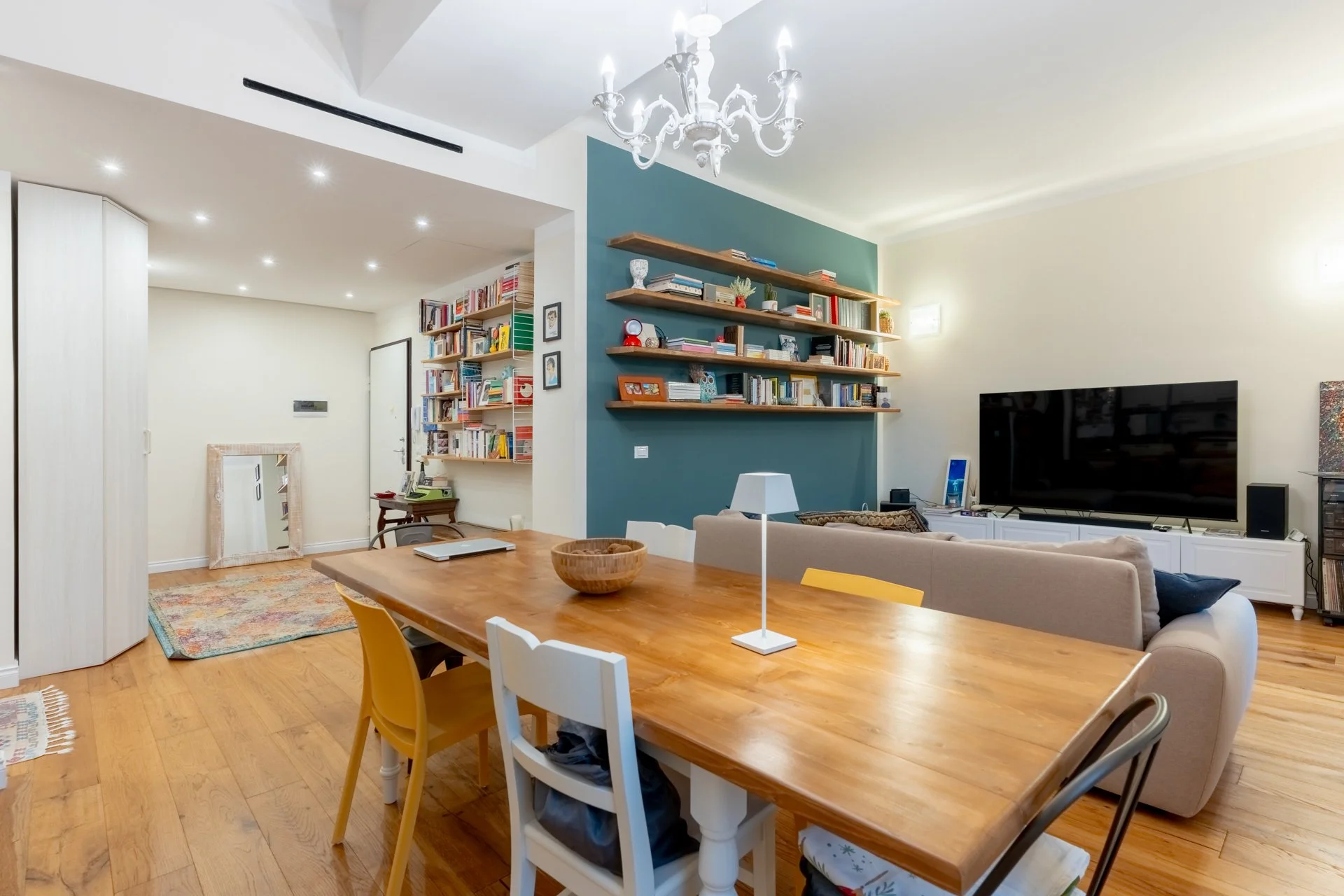 Soggiorno con parete blu, scaffali con libri, televisore, divano beige, tavolo in legno con sedie colorate, lampada da tavolo, pareti chiare, soffitto con lampadario bianco.