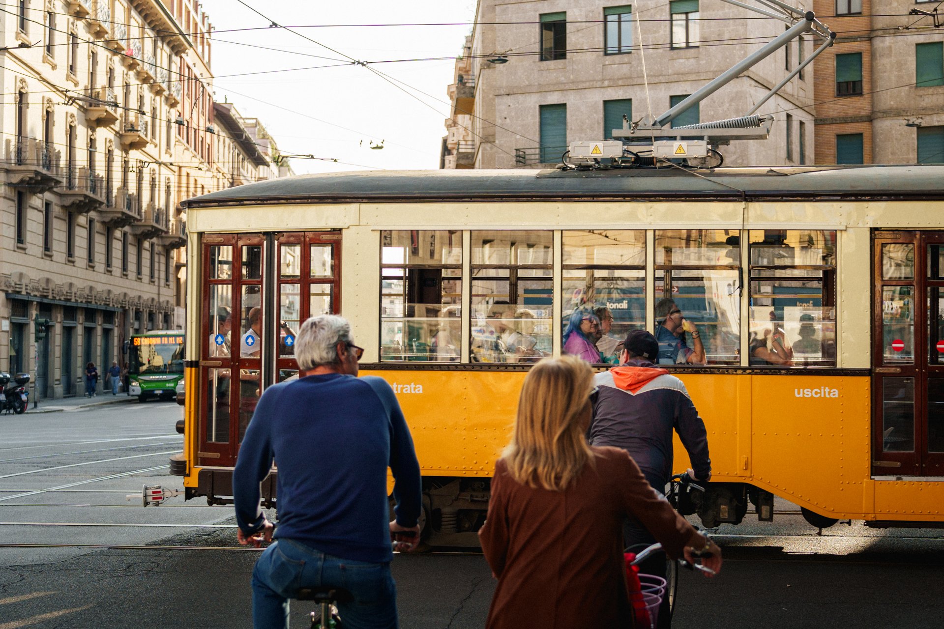 Una scena urbana con un tram giallo antico in movimento, passeggeri dentro e persone in bicicletta e pedoni sulla strada. Edifici storici e linee elettriche sopra la strada.