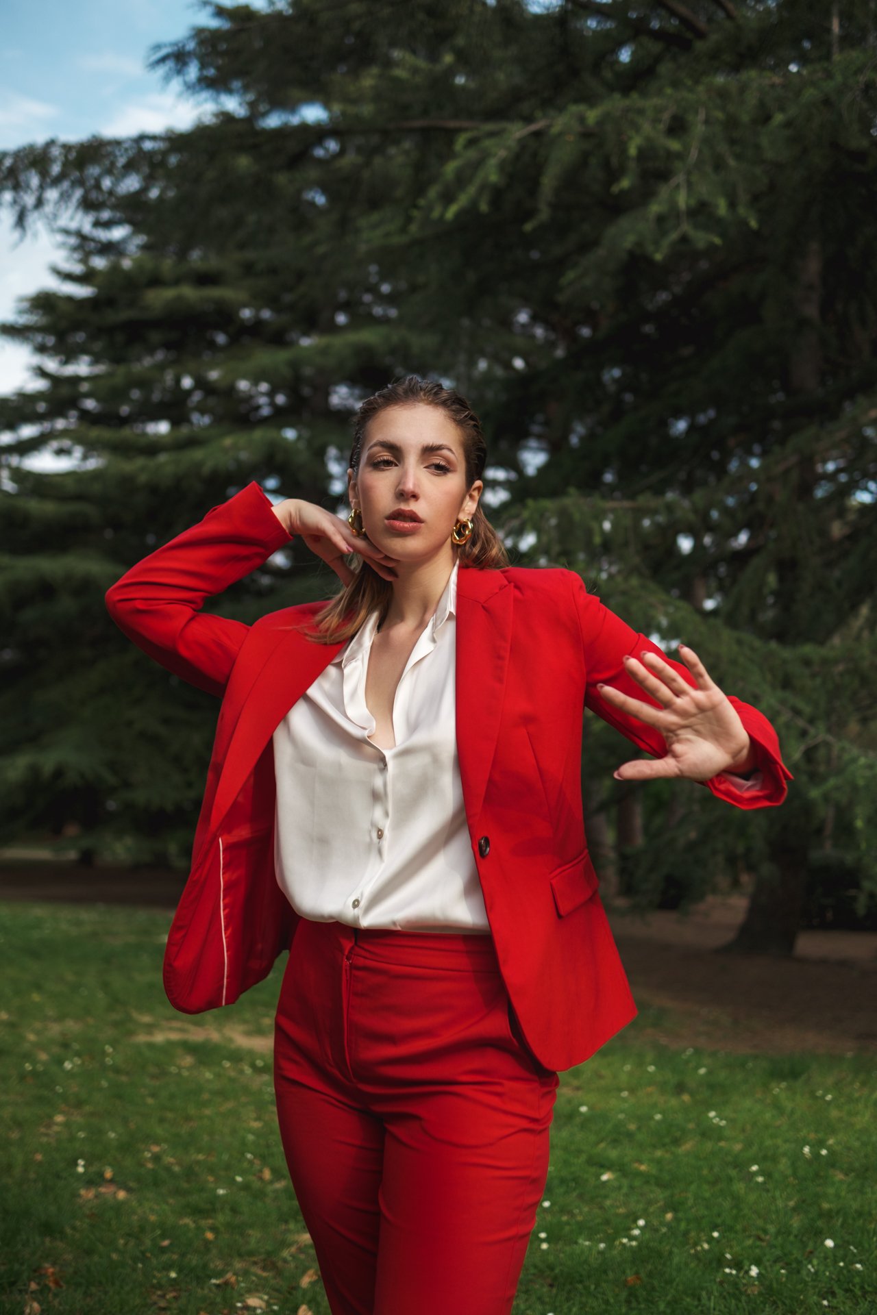 Una donna indossa un completo rosso con giacca e pantaloni, camicia bianca, e occhiali dorati, mentre si trova in un ambiente all'aperto con albero e cielo sullo sfondo.