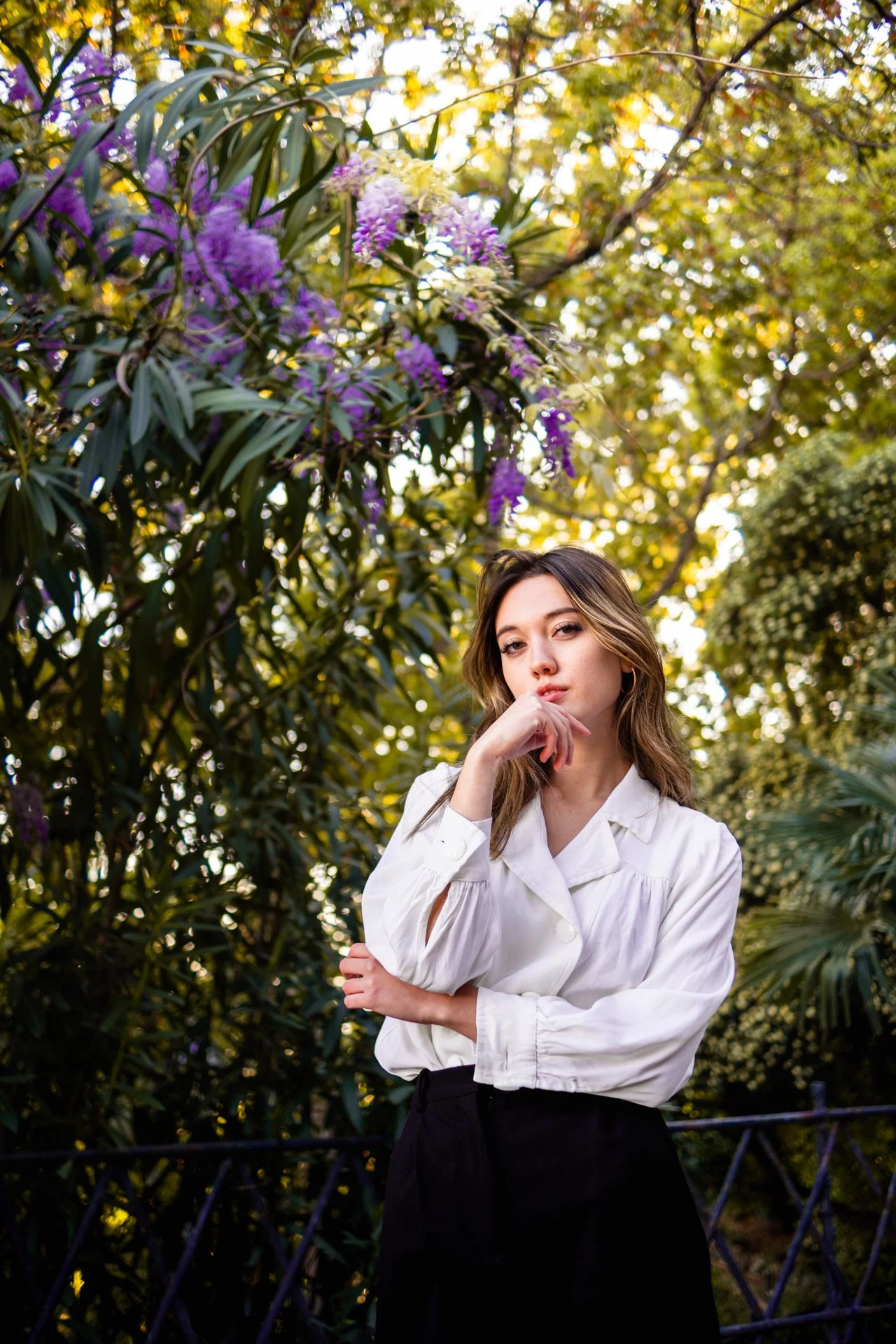 Una donna dai capelli castani con occhi chiari, vestita con una camicia bianca a maniche lunghe e una gonna nera, si trova in un giardino circondata da piante verdi e fiori viola e bianchi, con alberi e foglie dorate sullo sfondo.