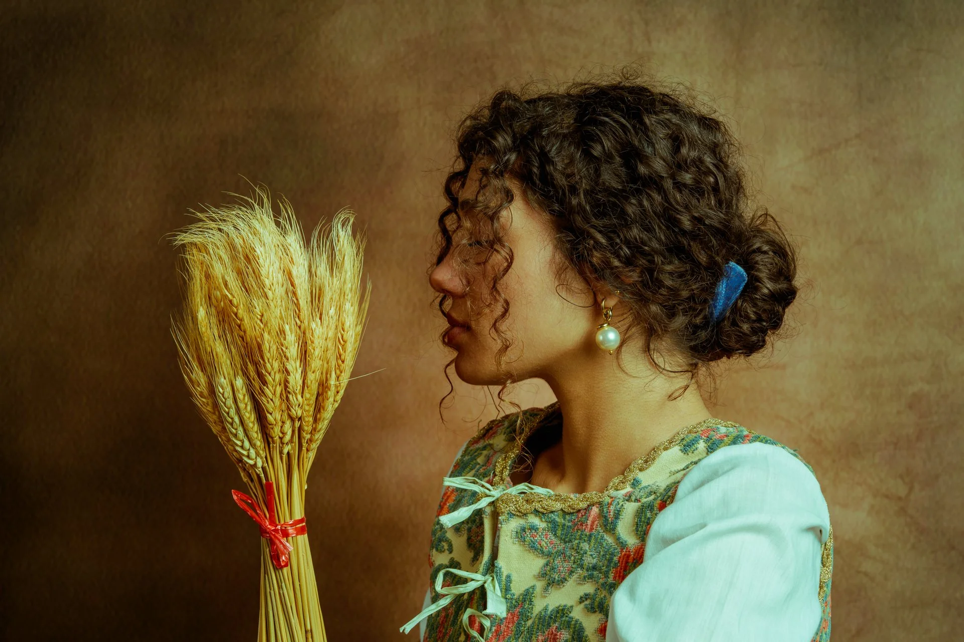 Ragazza con capelli riccioli e orecchino a perla guarda un fascio di grano legato con un tozzo di nastro rosso, sfondo marrone
