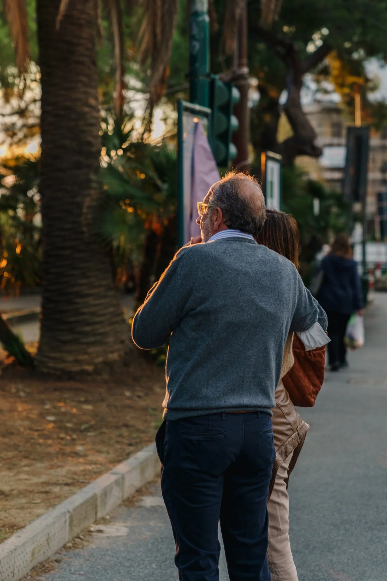 Uomo anziano con occhiali e giacca grigia che aspetta il semaforo rosso mentre si trova su un marciapiede con altre persone in una città
