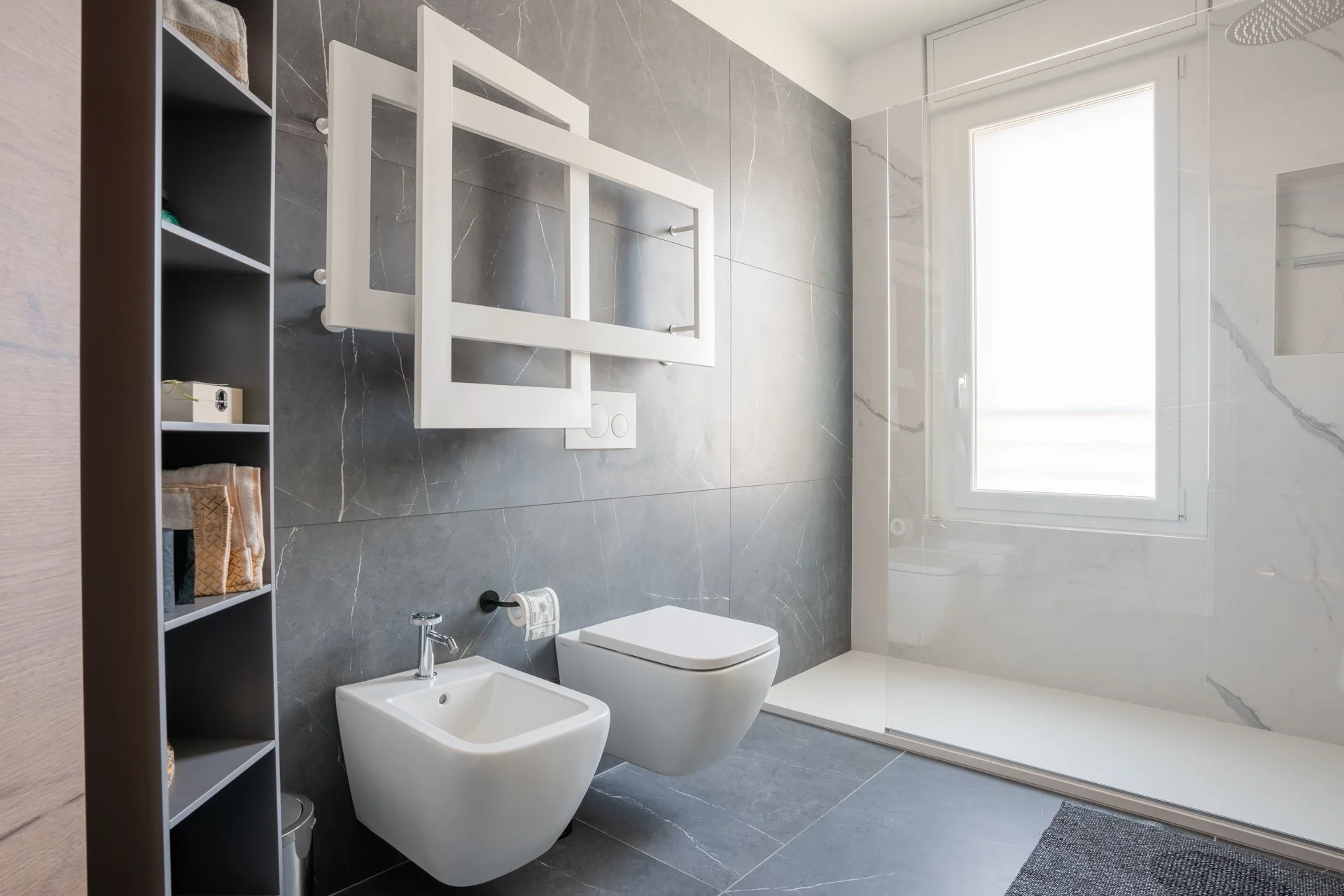 Bagno moderno con pareti in gres porcellanato grigio e bianco, lavabo, WC sospeso, bidet, grande finestra illuminata, e doccia a pavimento con vetri trasparenti.