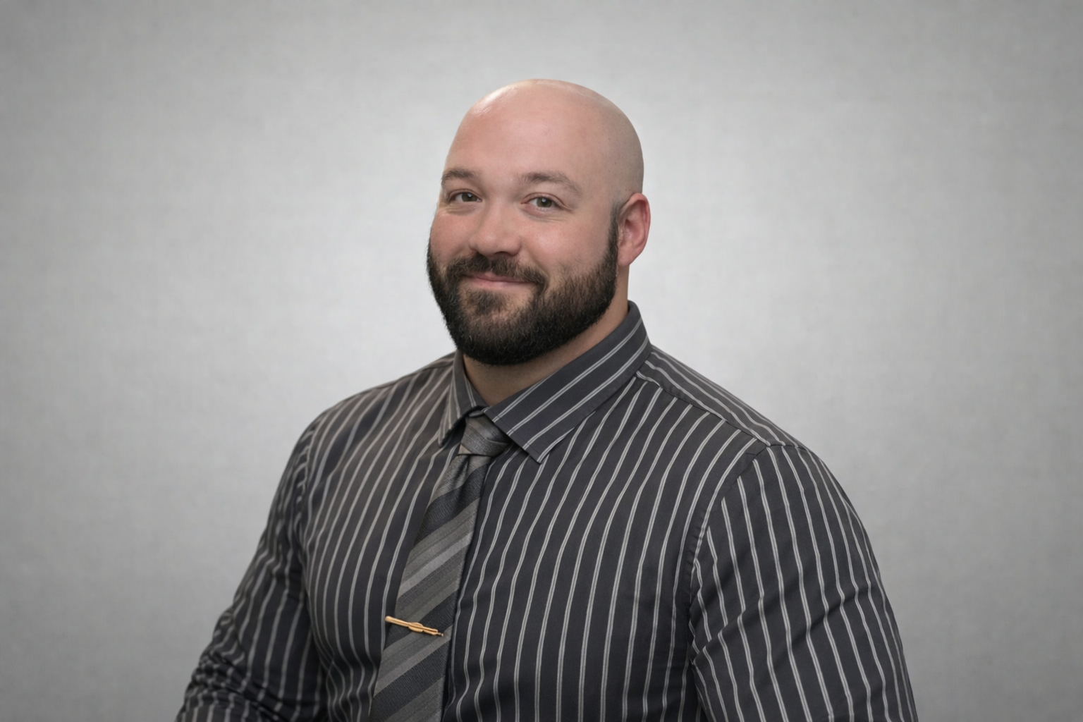 A man with a bald head, beard, and mustache wearing a dark gray pinstripe shirt and a gray tie, smiling slightly, against a plain light gray background.