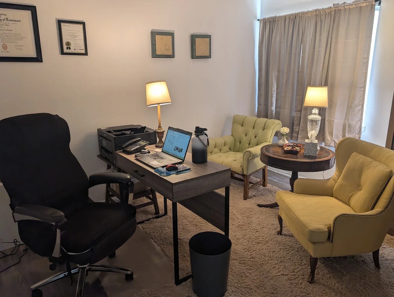 Office space with a desk, black office chair, laptop, desk lamp, and framed certificates on the wall. There are yellow armchairs, a round wooden table with a lamp and decorations, and curtains covering a window.