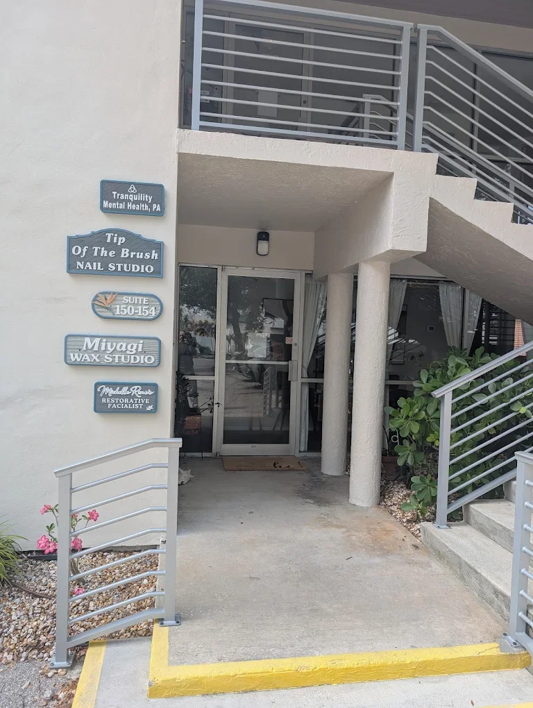 Entrance of a building with signs for nail studio, eyelash studio, and mental health services, with a stairway and plants nearby.