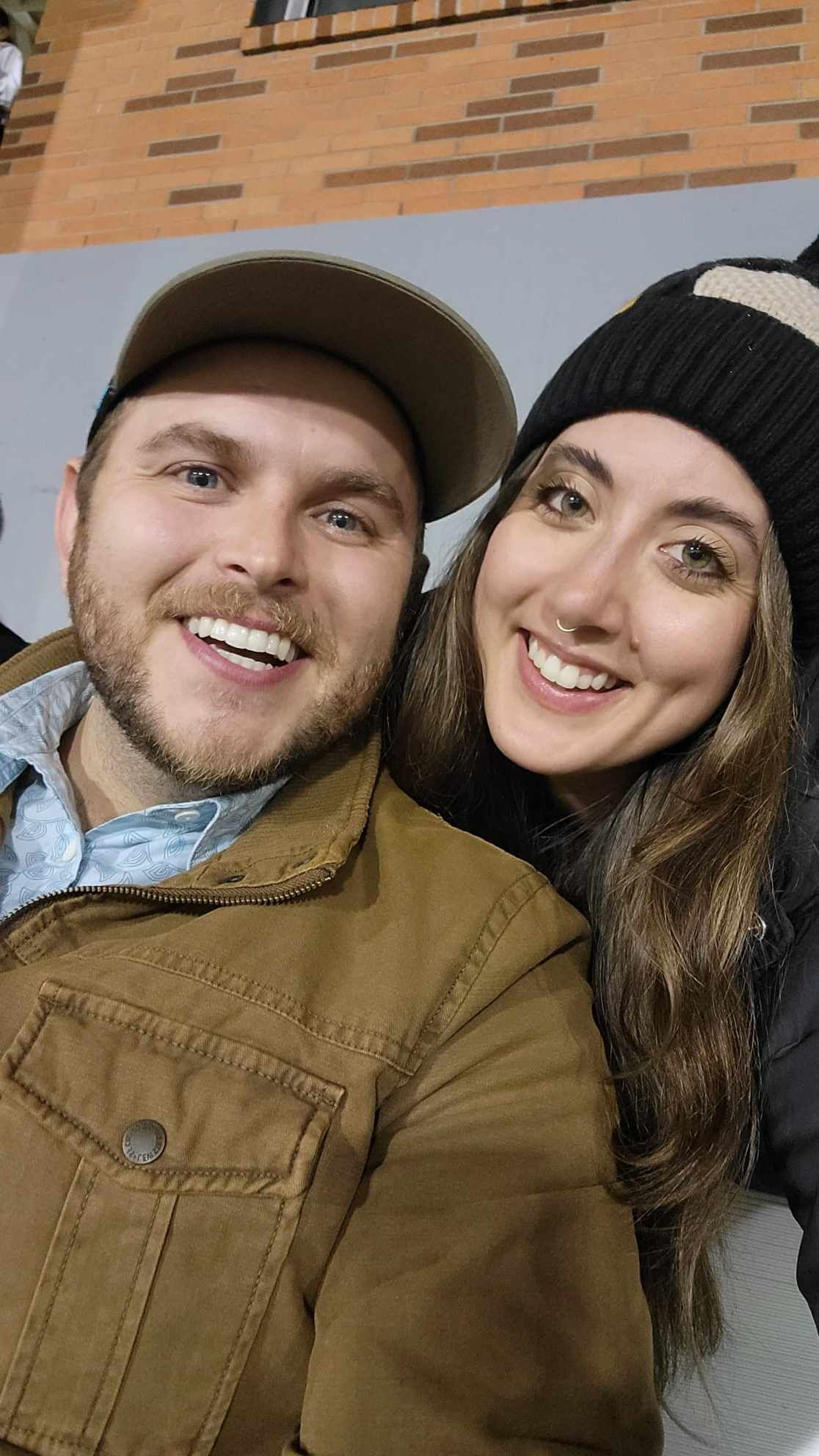 A smiling man with a beard and a baseball cap taking a selfie with a smiling woman wearing a black beanie and a septum piercing, in front of a brick wall.