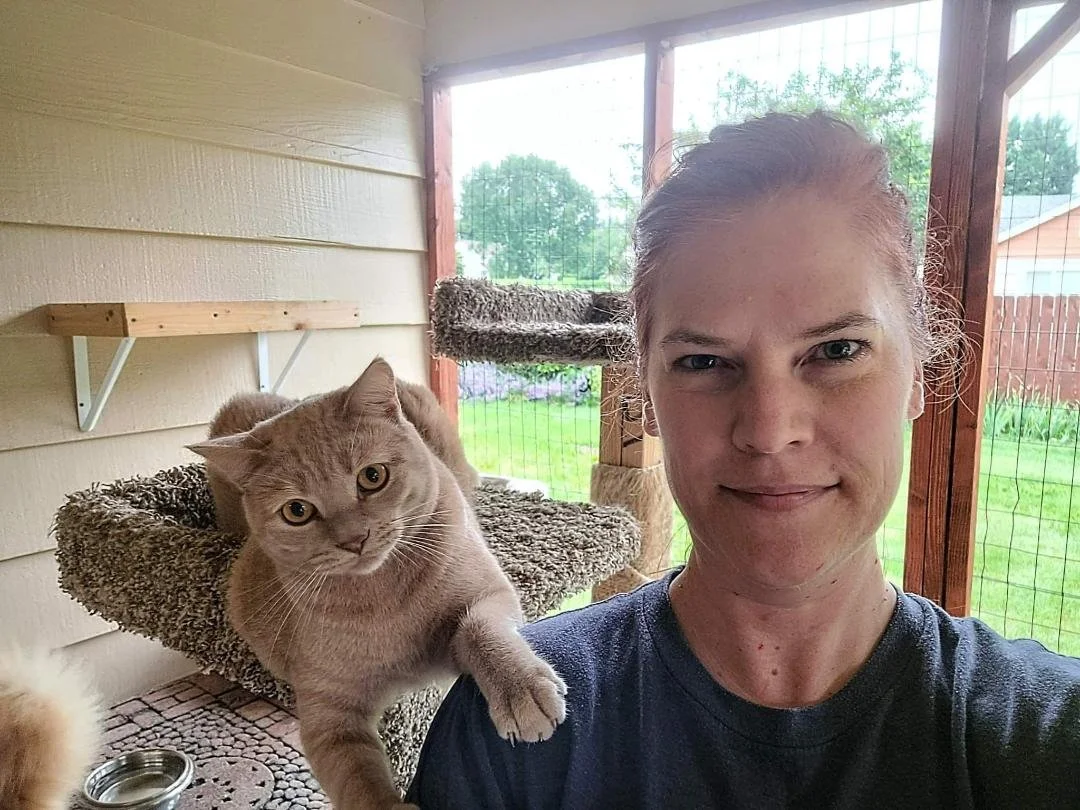 A woman taking a selfie with a gray cat on her shoulder inside a screened porch, with greenery visible outside.