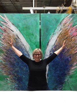 Person standing with arms raised in front of a large colorful abstract mural resembling butterfly wings.