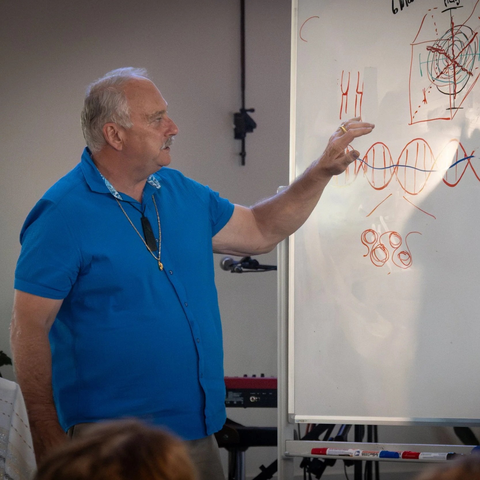 An older man with gray hair and a mustache, wearing a blue short-sleeved shirt, is giving a presentation or lecture while pointing at a whiteboard filled with colorful sketches and diagrams, including wavy lines, circles, and geometric shapes.