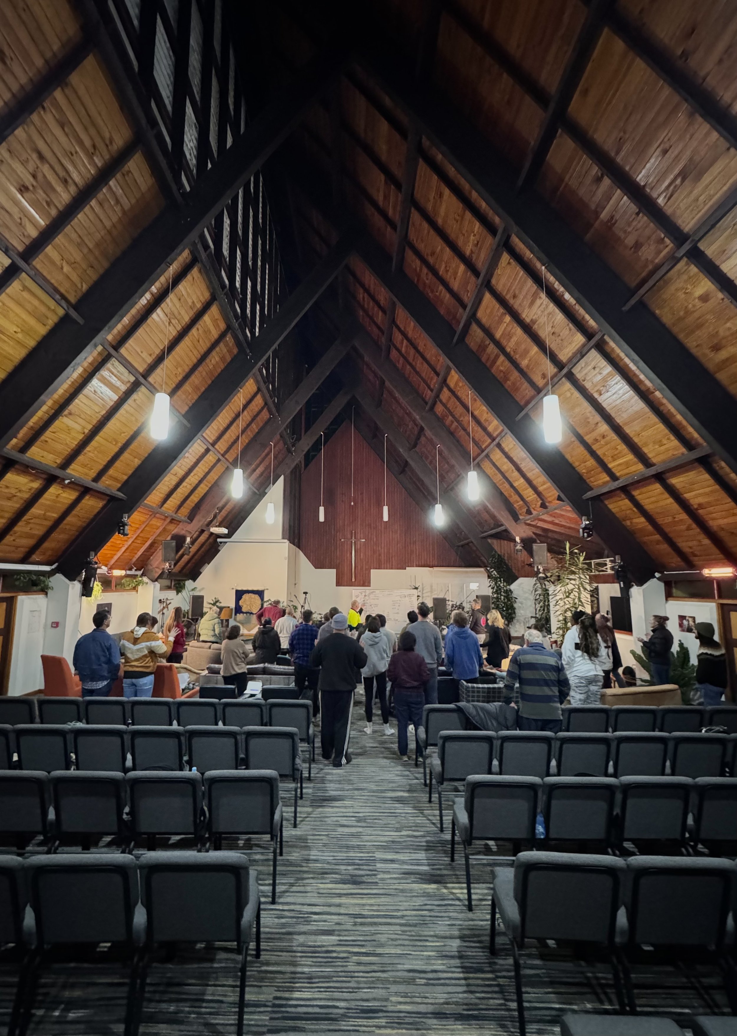 People gathered in a church with a wooden high ceiling, a cross on the wall, and rows of chairs.