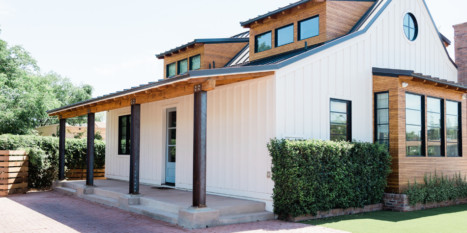 Modern two-story house with a white exterior, large windows, and a wooden upper section. It has a small front porch with four dark wooden posts, concrete steps, and nearby greenery.