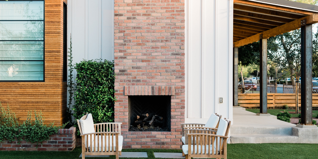 Outdoor seating with two wooden chairs with white cushions placed in front of a brick fireplace on a lawn, part of a modern house with wooden and white exterior walls and a covered patio area.