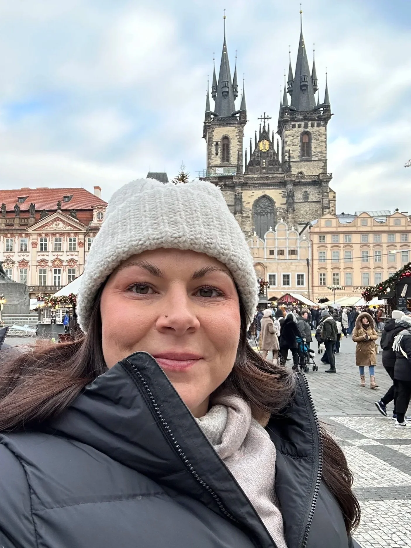 An anxiety therapist wearing a beige knit hat and black jacket takes a selfie in front of a busy square with a historic church in the background during winter.
