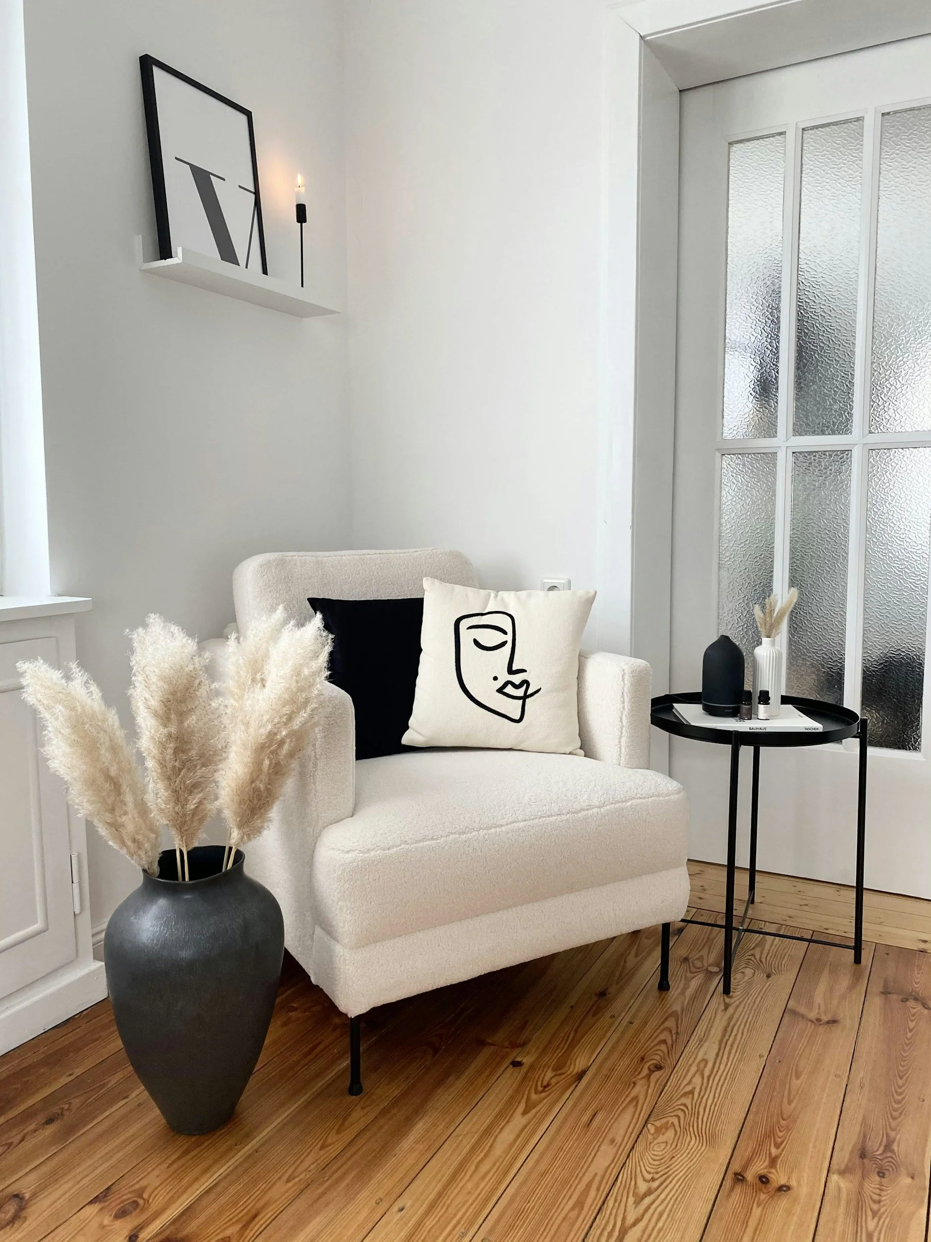 A cozy, modern living room corner featuring a white plush armchair with two decorative pillows, one black and one with a minimalist abstract face design. Next to the chair is a round black side table with vases and pampas grass. Behind the chair is a white wall with a floating shelf holding a framed artwork and a candle. The floor is wooden, and there is a frosted glass door nearby.