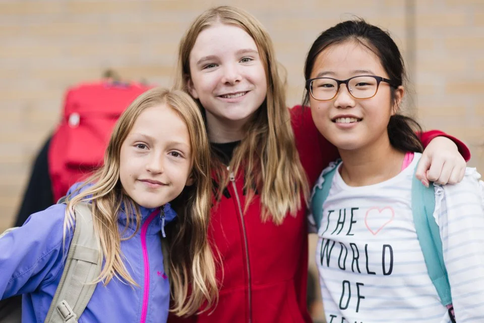 Three students smiling together outside a school building, showing friendship and community in a supportive learning environment.