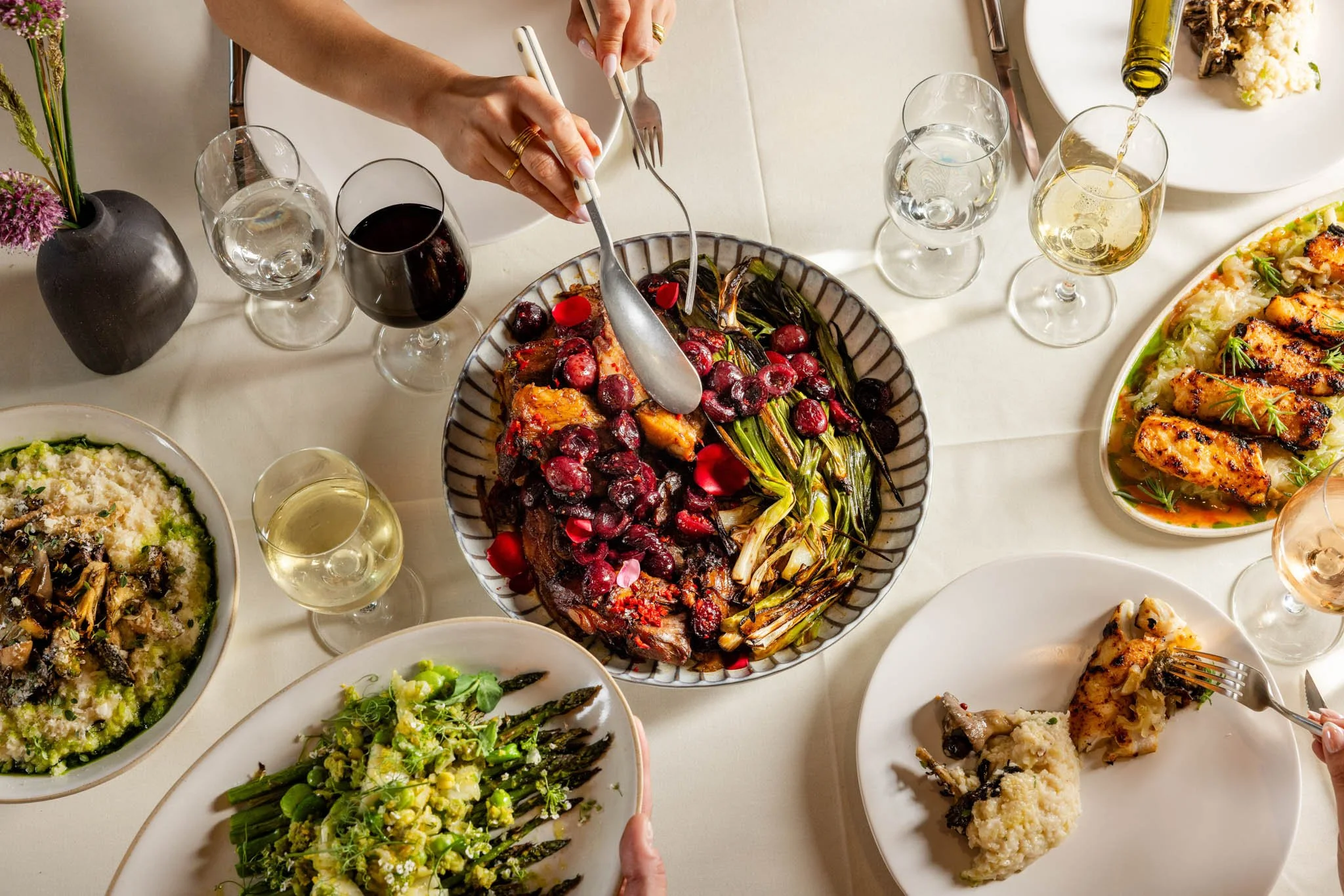 A dinner table with various dishes, including roasted vegetables, chicken, and asparagus, and several glasses of wine and water.
