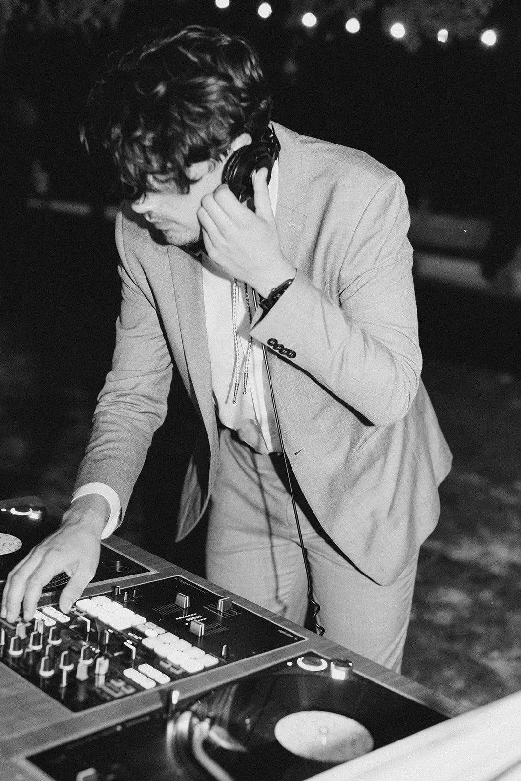 A man in a suit DJing at an event, wearing headphones, adjusting equipment on a DJ console at night.