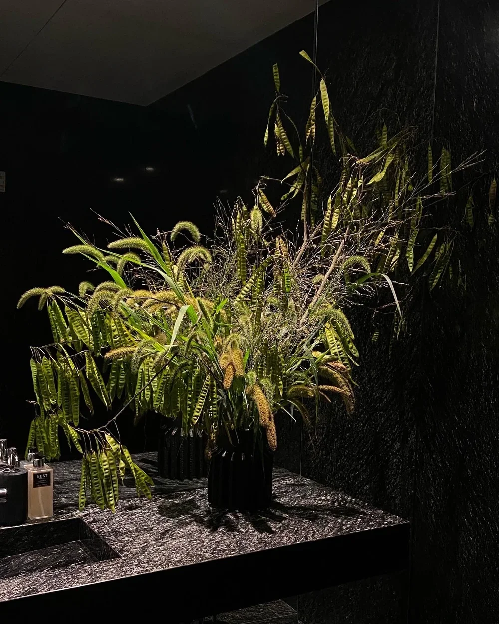 A large arrangement of green foliage and plant stalks in a black vase on a black textured countertop, with a dark wall background and toiletries to the left.